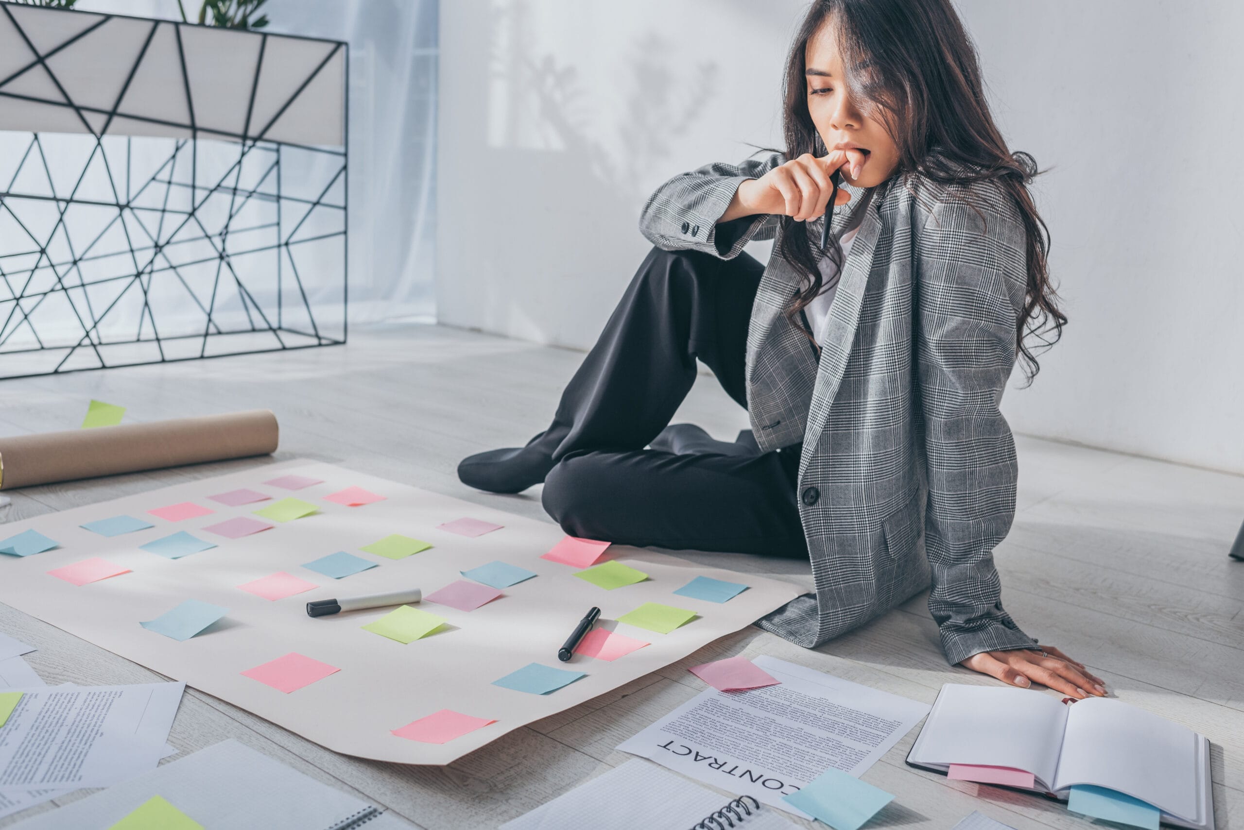 attractive asian scrum master sitting on floor thinking in office Acceptance Path Counseling A woman in a grey blazer sits thoughtfully on the floor surrounded by sticky notes and documents Strategic Implementation Your Clinical Intake and Roadmap