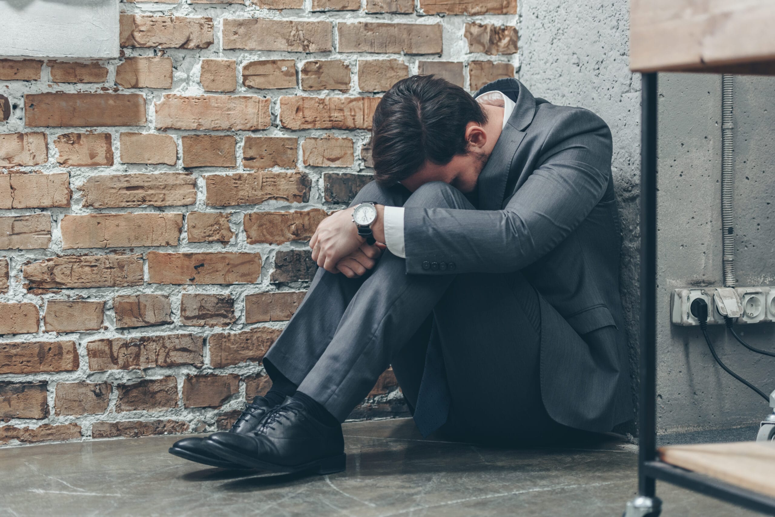 A man in a grey business suit sits huddled on the floor against a brick wall with his head bowed to his knees, his posture conveying a sense of profound distress and isolation.